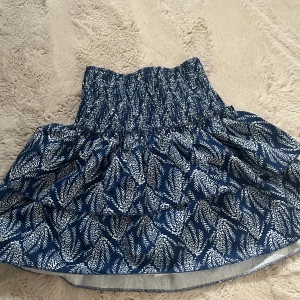 Blå mönstrad kjol med hög midja - Säljer en blå kjol med vitt mönster och hög smockad midja. De har ett luftigt material som är skönt en varm dag. Inga stora defekter endast några hängande trådar men inget man tänker på. Skulle säga XS men är stretchig i midjan