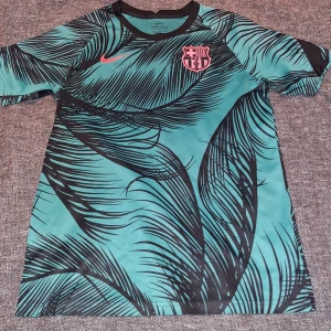 Barça Dri-Fit fotbollströja Nike L - Barcelona Dri-Fit fotbollströja från Nike i storlek L. Tröjan är turkos med svarta palmbladsmönster, har korta ärmar och klubbmärke samt Nike-logga i rosa på bröstet. Tillverkad i lätt och ventilerande polyester för maximal komfort på planen.