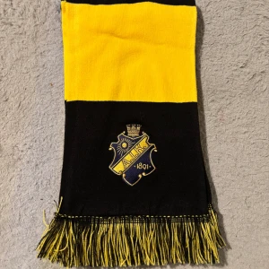 AIK halsduk svart och gul randig - Supporterhalsduk från AIK i klassiska svarta och gula breda ränder. Halsduken har broderat AIK-emblem och fransar i ändarna. Perfekt accessoar för att visa ditt lagstöd på matchen eller i vardagen.