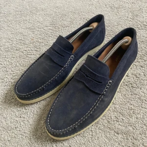 Mörkblå mockaloafers  - Snygga mörkblå loafers i mocka med kontrasterande vit sula och dekorativa sömmar runt tån. Klassisk penny loafer-design med rund tå och diskret detalj på ovandelen. Perfekta för en stilren och avslappnad look. Storlek 45 och sparsamt använda.