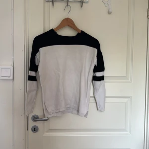 Svartvit stickad tröja från Rut&Circle - Snygg stickad tröja från Rut&Circle i svart och vitt. Tröjan har lång ärm, svart parti över axlar och armar samt vita partier med svarta ränder på ärmarna. Perfekt för dig som gillar enkel men stilren design.