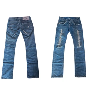 True Religion blå slitna jeans - Blå jeans från True Religion med slitna detaljer framtill och klassiska stora bakfickor med vit brodering. Jeansen har raka ben och låg midja.  Innerbenslängd -79-80cm Midja tvärs över - 39-40cm Unisex
