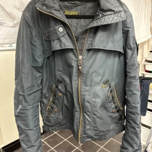 Mörkgrå vindjacka från Wiggy's - Snygg mörkgrå vindjacka från Wiggy's med huva, flera fickor med knappar och dragkedjor, samt coola detaljer på ärmen. Jackan har Lamalite-isolering och är perfekt för dig som gillar funktion och stil. Passar dig som vill ha en jacka med edge och streetkänsla.