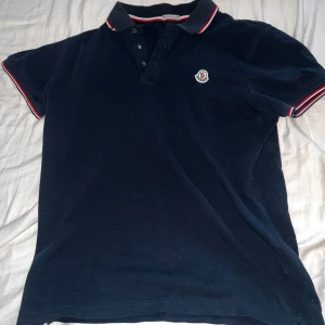 Marinblå pikétröja från Moncler - Snygg marinblå pikétröja från Moncler med klassisk krage och knappar framtill. Tröjan har röda och vita ränder på krage och ärmslut samt Moncler-logga på bröstet. Tillverkad i mjuk bomull som är skön mot huden. Perfekt för dig som gillar stilrena och sportiga vibes.