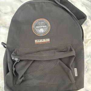 Svart ryggsäck från Napapijri - Snygg svart ryggsäck från Napapijri med klassisk logga framtill och en stor ytterficka med dragkedja. Perfekt storlek för skola eller häng, med justerbara axelremmar och slitstarkt material. Enkel och stilren design som funkar till det mesta.