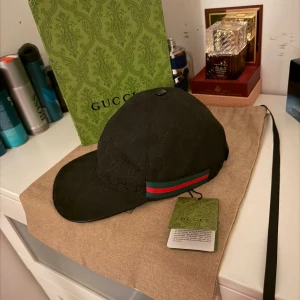 Svart Gucci keps - Svart keps från Gucci med diskret GG-monogram över hela kepsen och klassiska gröna och röda ränder på sidan. Justerbar rem baktill i skinn och detaljer i guld. Tillverkad i bomull och skinn, perfekt för dig som vill ha en lyxig och stilren accessoar.