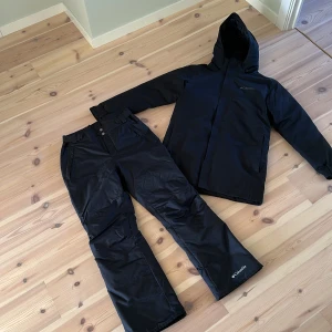 Svart jacka och byxa från Columbia för skidåkning/vinter - Svart vinterjacka från Columbia med huva och dragkedja, perfekt för kalla dagar. Jackan är fodrad och har en stilren look med diskret logga på bröstet. Materialet är slitstark polyester och jackan har långa ärmar och praktiska fickor. Med tillhörande byxor i boot cut fit. Finns en liten lagning på vänsterbenet som man knappt tänker på. 