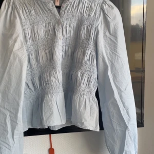 Ljusblå blus med smock från Nelly - Säljer en ljusblå blus från Nelly i storlek 38. Blusen har smockad överdel, knappar framtill och långa ärmar med lätt puff. Tillverkad i mjuk bomull som ger en skön och luftig känsla. Perfekt för dig som gillar romantisk och trendig stil.