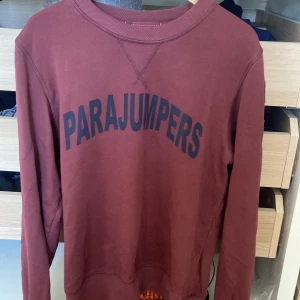 Vinröd fleece-tröja från Parajumpers - Säljer en vinröd fleece-tröja från Parajumpers med stort svart tryck på bröstet och patch på ärmen. Tröjan har rund hals, ribbade muddar och är långärmad. Perfekt för dig som gillar streetwear och vill ha något bekvämt och snyggt.