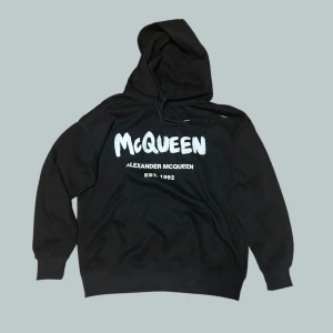 Alexander mqueen hoodie  - Svart hoodie från Alexander McQueen med stor vit logga och text på bröstet. Klassisk huva med snörning och känguruficka framtill. Perfekt för dig som gillar streetwear och vill sticka ut med en ikonisk design. Helt okej skick men lite använd.