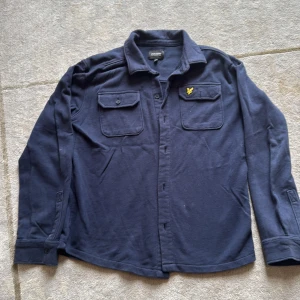 Mörkblå overshirt från Lyle & Scott - Snygg mörkblå overshirt från Lyle & Scott Junior med klassisk krage, två bröstfickor med knappar och gul broderad logga på ena fickan. Skjortan är tillverkad i mjuk bomull och har långa ärmar med knapp vid mudden. Perfekt för en chill och stilren look.