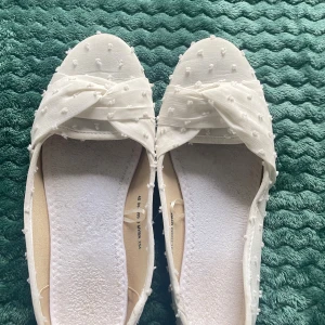 Vita ballerinaskor med rosett och prickar - Säljer ett par vita ballerinaskor med spetsig tå och dekorativ rosett framtill. Skorna har ett prickigt mönster med små tygprickar över hela ytan och är tillverkade i ett lätt tygmaterial. Perfekta för dig som gillar söta och stilrena detaljer. Har använt en gång under studenten. Är storlek 40 men har lagt in en sak så de blir 39 💌