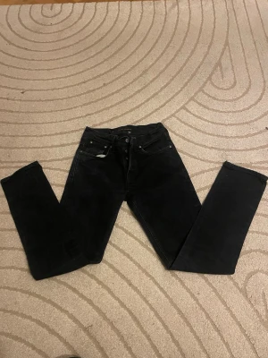 Svarta jeans från Nudie Jeans, 31/32 - Säljer ett par svarta jeans från Nudie Jeans i storlek 31/32. Klassisk femficksmodell med raka ben och diskret logga på bakfickan. Jeansen har en snygg, mörk tvätt och är tillverkade i slitstarkt bomullsmaterial. Perfekta för dig som gillar stilrena och tidlösa jeans.