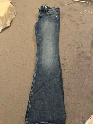 Blå bootcut jeans med låg midja - Säljer ett par klassiska blå jeans med bootcut-modell och låg midja. Jeansen har fem fickor, dragkedja och knapp framtill samt snygga slitningar för en avslappnad look. Perfekta till sneakers eller boots.