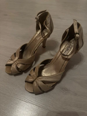 Guldiga pumps med öppen tå - Snygga pumps i satin från Vox Shoes. Skorna har öppen tå, smal klack och justerbart ankelband med spänne. De har ett elegant flätat mönster framtill och passar perfekt till dig som vill sticka ut lite extra.