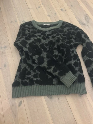 Grön leopardmönstrad stickad tröja - Cool stickad tröja i grönt med svart leopardmönster. Tröjan har rund halsringning och ribbstickade muddar vid ärmslut och nederkant. Perfekt för dig som gillar djurmönster och vill ha en trendig look.