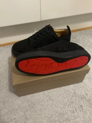 Christian Louboutin - Snygga svarta sneakers från Christian Louboutin i mocka med klassisk röd sula och svarta skosnören. Skorna har en låg profil och rund tå, samt beige innerfoder. Perfekta för dig som vill ha något stilrent med en lyxig touch.