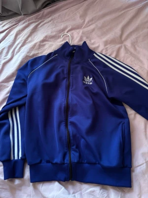 Blå adidas kofta - Säljer en klassisk blå ziptröja från Adidas med vita ränder längs ärmarna och Adidas-logga på bröstet. Tröjan har hög krage, dragkedja framtill och ribbade muddar. Perfekt för en sportig och avslappnad stil. 💙