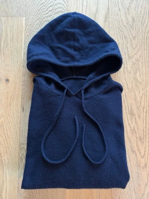 Mörkblå hoodie med huva i Kashmir - Mysig mörkblå hoodie i Kashmir med huva och dragsnören. Tillverkad i ett mjukt stickat material som känns skönt mot huden. Hoodien har långa ärmar och ribbade muddar vid ärmslut och nederkant. Perfekt för en avslappnad och chill stil. Från Snygg, svensk design.