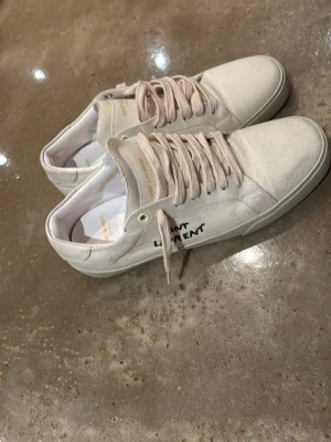 Saint Laurent vita sneakers i canvas - Säljer ett par stilrena vita sneakers från Saint Laurent i canvas med klassisk snörning och rund tå. Diskret Saint Laurent-logga på sidan och guldtext på plösen. Perfekta för en clean och avslappnad look.