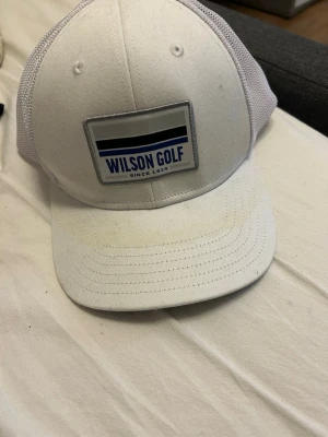 Vit Wilson Golf keps med mesh - Snygg vit keps från Wilson Golf med blå och svart logga framtill. Kepsen har klassisk böjd skärm och mesh-paneler bak för extra ventilation. Justerbar snapback gör att den passar de flesta. Perfekt för golfbanan eller som stilren accessoar.