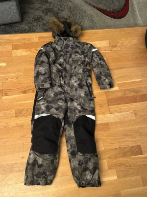 Camouflage täckbyxor med huva - Täckbyxor i camouflage-mönster med svart förstärkning på knän och bak. Utrustad med huva och fuskpäls, samt reflexdetaljer på ärm och ben. Perfekt för kalla vinterdagar och utomhusaktiviteter.