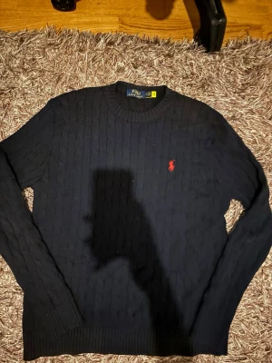 Mörkblå kabelstickad tröja Polo Ralph Lauren - Klassisk mörkblå kabelstickad tröja från Polo Ralph Lauren med rund halsringning och den ikoniska röda logotypen broderad på bröstet. Tröjan har långa ärmar och ribbade muddar vid ärmslut och nederkant. Perfekt för en stilren och tidlös look.
