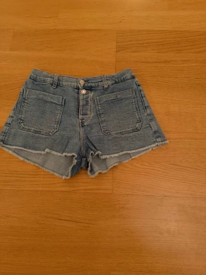 Blå jeansshorts med rå kant - Snygga blå jeansshorts med mid waist och rå, fransig kant. Shortsen har stora fickor både fram och bak samt dubbla knappar i midjan. Perfekta för sommaren och ger en avslappnad look.