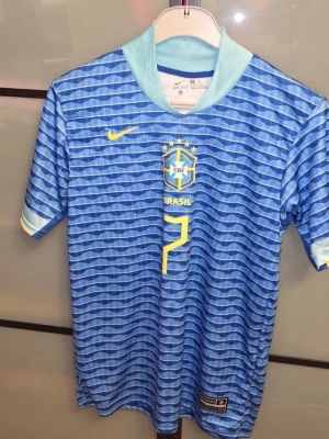 Blå Brasilien fotbollströja Nike - Snygg blå Brasilien fotbollströja från Nike med gult tryck, nummer 7 och VINI JR på ryggen. Tröjan har vågigt mönster, korta ärmar och ljusblå krage. Perfekt för dig som älskar fotboll och vill stötta landslaget. Den är helt ny, aldrig använd men den är tvättad. Tveka inte att ställa frågor! Priset är inte hugget i sten.