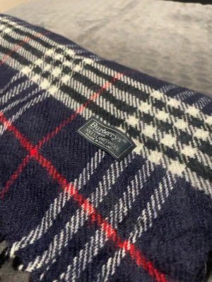 Mörkblå rutig halsduk från Burberry - Klassisk halsduk från Burberry i mörkblått med rutigt mönster i vitt och rött. Tillverkad i 100% lammull och har fransar i kanterna. Perfekt accessoar för att lyfta din outfit med en tidlös brittisk stil.