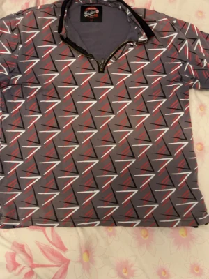 Grå mönstrad polotopp med dragkedja - Cool grå polotopp från Galatasaray med grafiskt mönster i svart, vitt och rött. Toppen har korta ärmar, halv dragkedja framtill och en sportig känsla. Perfekt för dig som gillar unika prints och vill sticka ut.