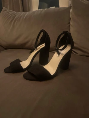 Svarta pumps med hög klack och ankelrem - Stilrena svarta pumps med bred hög klack och öppen tå. Skorna har en enkel ankelrem med spänne och är tillverkade i mocka-liknande material. Perfekta för dig som vill ha en klassisk men trendig look.