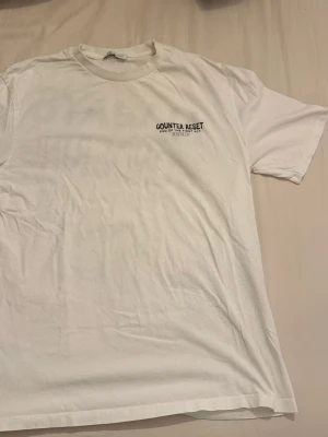 Vit Counter Reset t-shirt Zara  - Vit t-shirt från Counter Reset med svart tryck på bröstet. Klassisk rund hals och korta ärmar. Tillverkad i mjuk bomull för en skön och avslappnad stil. Perfekt till jeans eller shorts.
