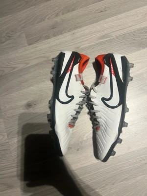 Nike vita fotbollsskor med svart swoosh - Säljer ett par snygga vita Nike fotbollsskor med svart swoosh och orange detaljer. Skorna har snörning och är designade för bra grepp på planen med fasta dobbar. Perfekta för dig som vill sticka ut på matchen!