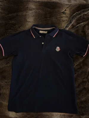 Marinblå pikétröja från Moncler - Snygg marinblå pikétröja från Moncler med klassisk krage och två knappar. Tröjan har vita och röda detaljer på krage och ärmslut samt Moncler-logga broderad på bröstet. Perfekt till jeans eller shorts för en clean look.