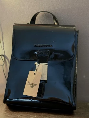 Svart ryggsäck från Dr. Martens - Snygg svart glansig ryggsäck från Dr. Martens. Klassisk fyrkantig form med justerbara axelband och ett praktiskt handtag upptill. Perfekt för dig som vill ha en stilren och edgy accessoar till vardagen. Helt ny och orörd. Nypris var 1400 kr. 