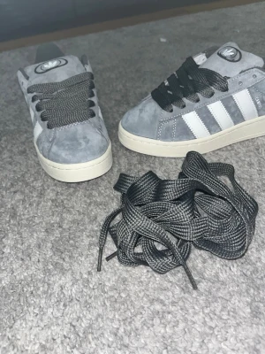 Adidas Campus grå sneakers mocka helt nya  - Grå Adidas Campus sneakers i mocka med vita klassiska ränder på sidorna och vit sula. Skorna har snörning och låg profil, samt extra skosnören. Perfekta för en stilren och avslappnad look.