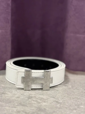 Vitt läderbälte med H-spänne - Snyggt vitt bälte i läder med ett stort, silverfärgat H-format metallspänne. Perfekt accessoar för att lyfta din outfit och ge en exklusiv touch. Bältet har flera hål för justerbar passform och en stilren design.