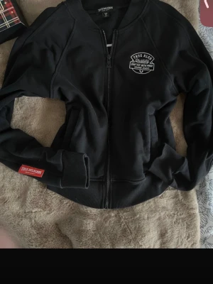 Svart zip-hoodie från True Religion - Svart hoodie med dragkedja från True Religion. Stor vit logga på ryggen och mindre logga på bröstet. Ribbade muddar och midja, samt True Religion-märke på ärmen. Skön och stilren modell i mjukt material, perfekt till jeans eller joggers.