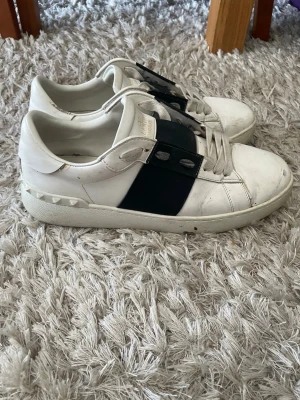 Valentino Garavani opens svarta - Snygga vita sneakers från Valentino Garavani med svart band över vristen och ikoniska nitar på hälen. Skorna har snörning och är tillverkade i skinn för en exklusiv känsla. 8/10 skick, ser värre ut än de är då dom är ganska smutsiga. Kom med frågor o prisförslag 