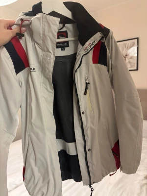 Vit vindjacka från Marine - Snygg vit vindjacka från Marine med marinblå och röda detaljer. Jackan har dragkedja framtill, meshfoder och justerbar huva. Perfekt för blåsiga dagar och har en sportig look med logga på bröstet.