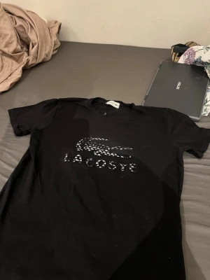 Svart Lacoste t-shirt S-M - Snygg svart t-shirt från Lacoste med det klassiska krokodilloggan och texten på bröstet i glansigt material. T-shirten har rund halsringning och korta ärmar, perfekt för en stilren och sportig look.
