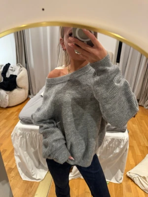 Oversized grå stickad tröja - Mysig oversized stickad tröja i ljusgrått med bred ribbstickning och nedhasad axel. Tröjan har långa ärmar och en avslappnad passform, perfekt för lager-på-lager och chill vibes. Materialet ser ut att vara mjukt och stretchigt, vilket gör den extra bekväm.