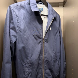 Marinblå vindjacka från Polo Ralph Lauren - Snygg marinblå vindjacka från Polo Ralph Lauren med klassisk krage och dragkedja framtill. Jackan har diskret logga på bröstet och två sidofickor. Perfekt för dig som vill ha en stilren och enkel jacka till vardags.
