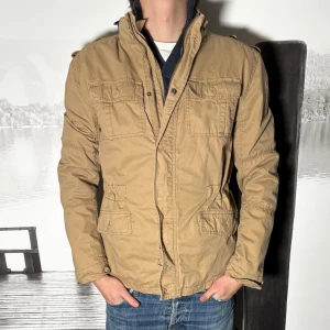 Beige vindjacka med fickor - Snygg beige vindjacka med fyra framfickor och hög krage. Jackan har dragkedja framtill och knappdetaljer på fickorna. Perfekt för dig som vill ha en stilren och funktionell jacka till vardags. Passformen är normal och jackan har långa ärmar.