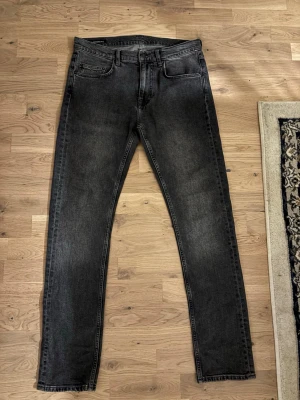 Grå/svarta jeans från J.Lindeberg - Säljer ett par snygga grå/svarta jeans från J.Lindeberg med klassisk femficksdesign och diskret logga på myntfickan. Jeansen har rak passform och är tillverkade i slitstarkt denimtyg. Perfekta för dig som gillar stilrena plagg. Jeansen är nyskick och knappt använda mer än ett par gånger.