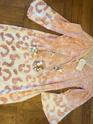 Långklänning med rosa leopardmönster - Trendig långklänning med rosa leopardmönster och vida ärmar. Klänningen har v-ringning och dekorativa snören med pärlor och tofsar framtill. Tillverkad i ett mjukt, lätt material som passar perfekt för varmare dagar.