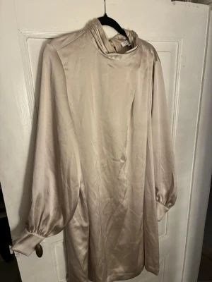 Champagnefärgad sidenklänning Stockh LM - Elegant champagnefärgad klänning från Stockh LM i glansigt sidenmaterial. Klänningen har lång ärm med bred manschett och hög krage med draperad effekt baktill. Perfekt för dig som gillar stilrena och lyxiga plagg.