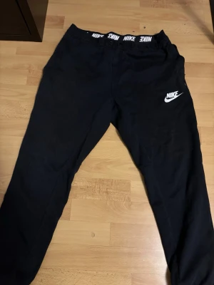 Svarta mjukisbyxor från Nike - Svarta mjukisbyxor från Nike med vit logga på vänster ben och elastisk midja med Nike-tryck på insidan. Byxorna har en klassisk passform och är tillverkade i mjukt material som känns skönt mot huden. Perfekta för en avslappnad stil.