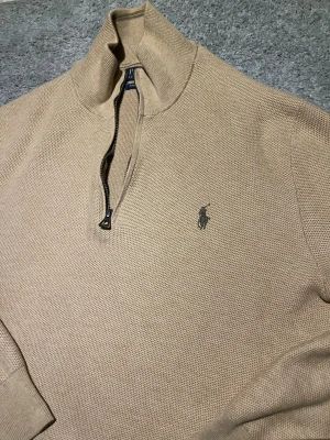 Beige halvzip-tröja från Polo Ralph Lauren - Klassisk beige stickad tröja från Polo Ralph Lauren med halv dragkedja och hög krage. Tröjan har det ikoniska broderade logotypen på bröstet och är tillverkad i ett grovt stickat material som ger en stilren och avslappnad look.